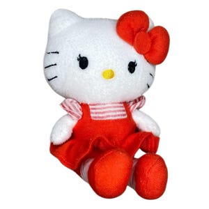 May include: A plush white cat doll with a red bow, dress, and shoes. The doll has black eyes, whiskers, and a yellow nose. The dress has red and white striped sleeves. The doll is a popular character.