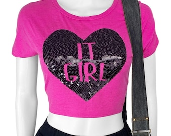 Puede incluir: Una camiseta corta rosa brillante con un diseño de corazón negro con lentejuelas. El corazón presenta las palabras "IT GIRL" en rosa. La camiseta tiene mangas cortas y cuello redondo. Se ve un bolso de hombro de mezclilla negro.