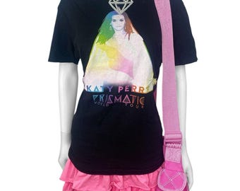 Puede incluir: Camiseta negra con un gráfico de "Prismatic World Tour" de Katy Perry, combinada con una falda rosa con volantes y un bolso bandolera rosa. Un collar con colgante de diamante plateado completa el conjunto. El maniquí está sobre un fondo blanco.