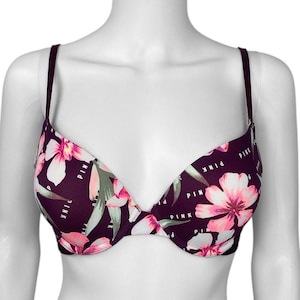 Puede incluir: Un sujetador push-up con estampado floral. El sujetador presenta un fondo morado oscuro con flores rosas y blancas y hojas verdes. La palabra "PINK" est&aacute; impresa en blanco en el sujetador.