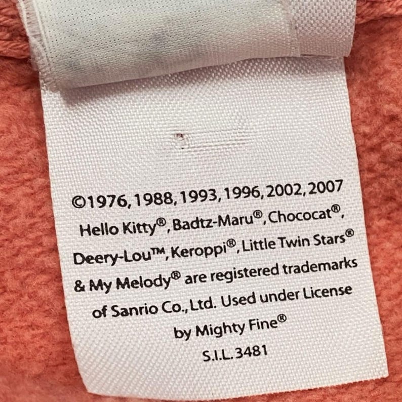 May include: A white fabric tag with black text, featuring the registered trademarks of Sanrio characters like Hello Kitty and My Melody. The tag also includes the years 1976, 1988, 1993, 1996, 2002, and 2007.
