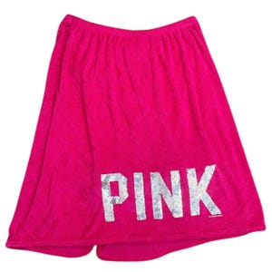 May include: A hot pink terry cloth wrap with the word "PINK" in silver sequined letters. The wrap appears to be new and unused. The wrap is designed to be worn around the waist.