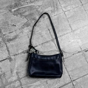 May include: A black leather shoulder bag with a long strap. The bag has a rectangular shape with a slightly curved top and a textured surface. The bag is sitting on a gray, textured surface.