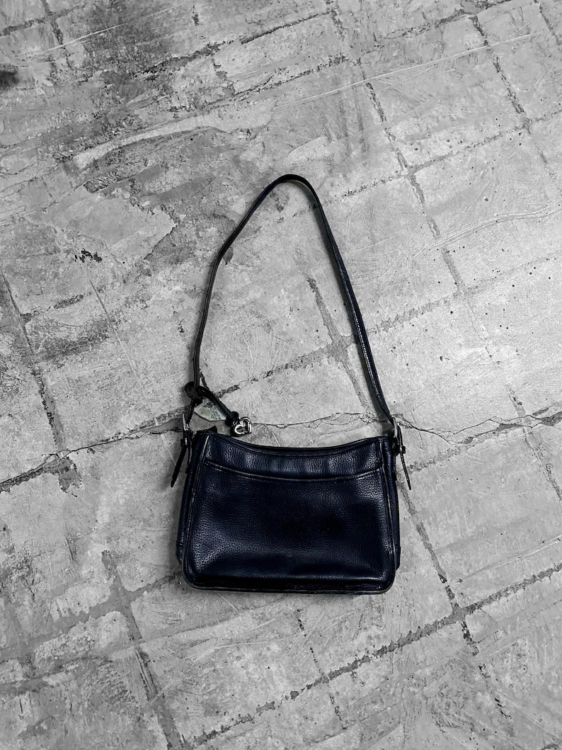 May include: A black leather shoulder bag with a long strap. The bag has a rectangular shape with a slightly curved top and a textured surface. The bag is sitting on a gray, textured surface.