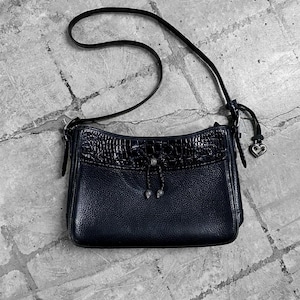 May include: A black leather crossbody bag with a textured flap and a long strap. The bag features a silver heart charm and a decorative tie detail. The bag is rectangular and has a pebbled texture.