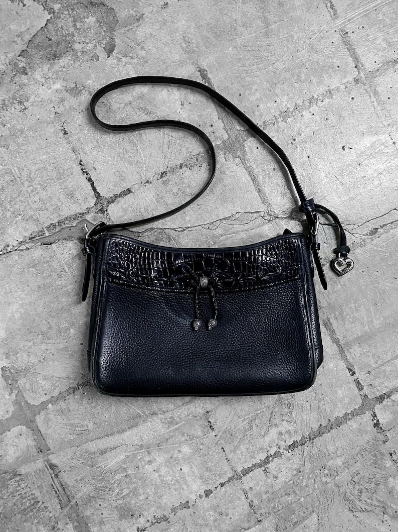 May include: A black leather crossbody bag with a textured flap and a long strap. The bag features a silver heart charm and a decorative tie detail. The bag is rectangular and has a pebbled texture.