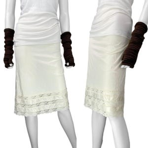 May include: Cream-colored slip skirt with a lace trim at the hem. The skirt is paired with brown fingerless gloves and a white tank top. The skirt is knee-length and has a satin-like texture.