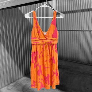 May include: An orange and pink floral print dress with a V-neck and pleated waist detail. The dress is sleeveless and hangs on a white hanger. The fabric has a soft, flowing appearance.