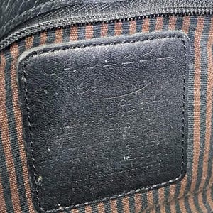 May include: Close-up of a black leather bag interior. The image shows a rectangular leather patch with a stitched border, set against a striped fabric lining in brown and black. A zipper is visible at the top.