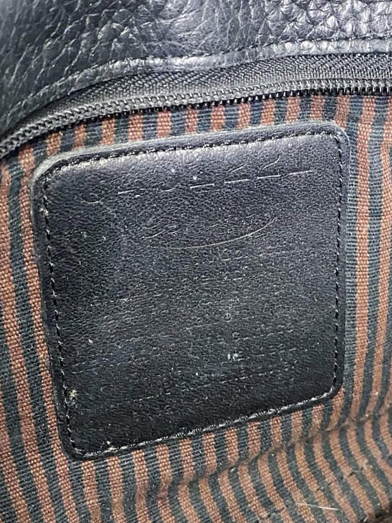May include: Close-up of a black leather bag interior. The image shows a rectangular leather patch with a stitched border, set against a striped fabric lining in brown and black. A zipper is visible at the top.