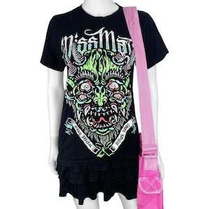 May include: Black t-shirt with a green, pink, and blue monster graphic and the text "Miss May". The shirt has a layered ruffle hem. A pink crossbody bag with a pocket is over the shoulder.