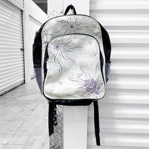 May include: A white and black backpack with a floral design and the text "Ak & Aj" in purple. The backpack has a front pocket, side mesh pockets, and black straps.