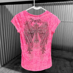 May include: A bright pink t-shirt with a rhinestone-studded cross and angel wings design on the back. The word "Faith" is arched above the cross. The shirt is hanging on a white hanger.