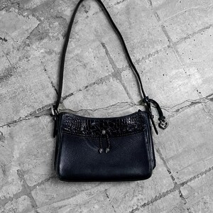 May include: A black leather shoulder bag with a textured crocodile-like flap. The bag has a long, thin strap and a small heart-shaped charm. The bag is set against a gray, textured background.