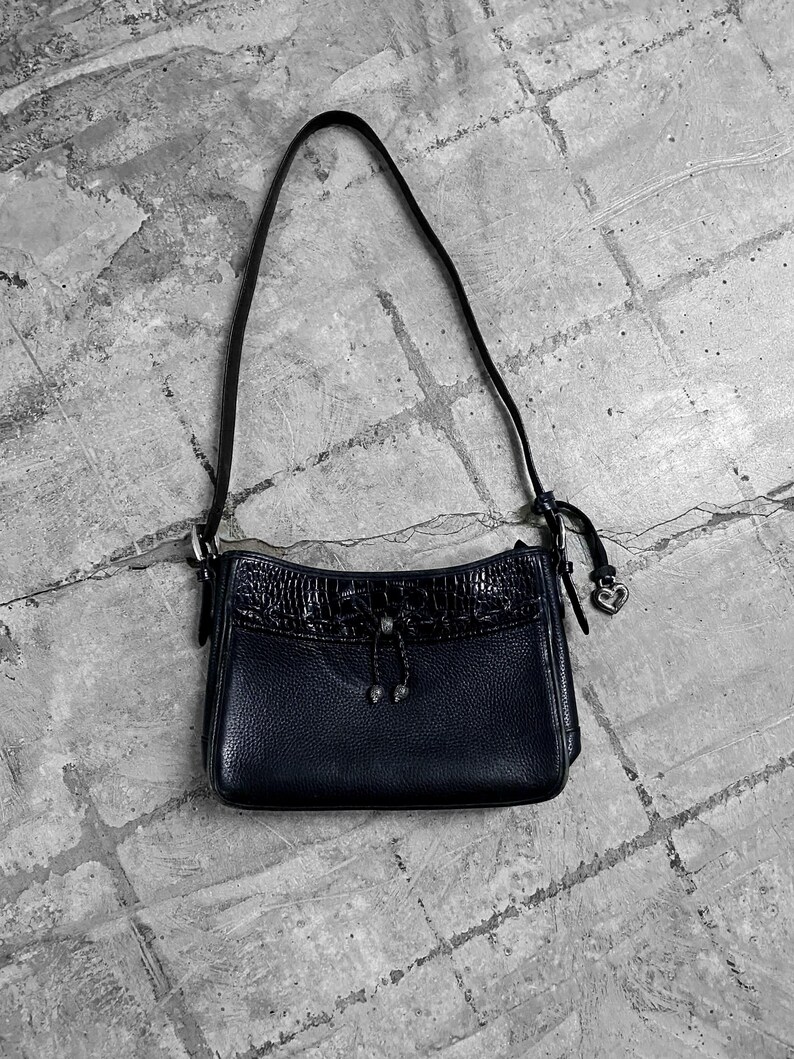 May include: A black leather shoulder bag with a textured crocodile-like flap. The bag has a long, thin strap and a small heart-shaped charm. The bag is set against a gray, textured background.