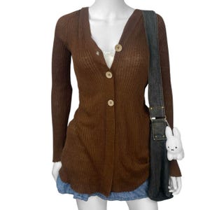 Puede incluir: Un cárdigan marrón de punto acanalado con cuello en V y cierre de botones. El suéter de manga larga se combina con una falda vaquera y un bolso de hombro de mezclilla oscura con un dije de conejo de peluche blanco.