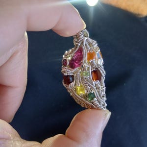 Peut inclure: Un pendentif artisanal avec un design en fil de fer. Le pendentif présente une mosaïque de pierres précieuses colorées, notamment rouges, roses, oranges, jaunes et vertes. Le fil est argenté, créant un contraste avec les pierres vibrantes. Le pendentif est tenu dans une main.