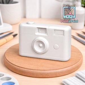 Puede incluir: Un objeto blanco con forma de cámara con lente, visor y botón, apoyado sobre una base circular de madera. El objeto es de color sólido y tiene un diseño retro. La imagen también incluye el texto "Print & Paint".