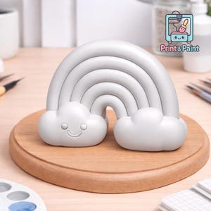 Könnte beinhalten: Ein hellgraues, regenbogenförmiges Deko-Objekt mit lächelnden Wolken an jedem Ende. Der Regenbogen steht auf einer runden Holzbasis. Das Bild enthält auch den Text "Print & Paint".