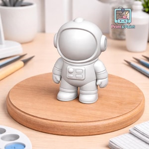 May include: A small, unpainted astronaut figurine stands on a wooden circle. The astronaut is gray and has a helmet, a rectangular chest panel, and rounded boots. The figurine is ready to be painted. The logo "Print & Paint" is visible in the background.
