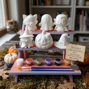 Op de afbeelding: Een houten displaystandaard met drie niveaus met zes witte, onbeschilderde beeldjes, waaronder een kat, geest, vampier, pompoen en heksenhoed. De set bevat zes kleine verfpotjes, twee blauwe penselen en een bordje met Spooky Creations Kit.