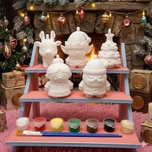 May include: A set of five white ceramic holiday figurines including a reindeer, an elf, a Christmas tree, Mrs. Claus, and Santa Claus, displayed on a three-tiered wooden stand with six small paint pots and two paintbrushes on the bottom shelf.
