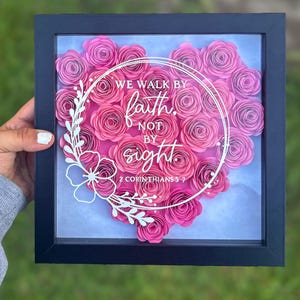 May include: A framed piece of art with a heart made of pink paper roses. White text within the heart reads "WE WALK BY faith, NOT BY sight" and "2 CORINTHIANS 5:7". The frame is black, and the artwork is square.