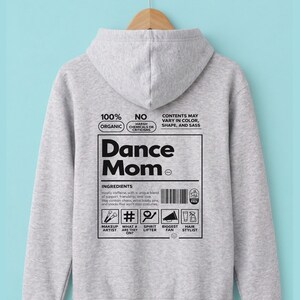 Puede incluir: Una sudadera con capucha gris claro con las palabras "Dance Mom" en negrita, en negro. La parte posterior de la sudadera con capucha presenta un gráfico con texto sobre ingredientes e iconos que representan maquillaje, espíritu y peluquería. La sudadera con capucha está colgada en una percha de madera.
