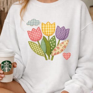 May include: White sweatshirt with a colourful tulip design. The tulips are pink, yellow, and purple, with green leaves and a small blue cloud. The design has a patchwork style with polka dots, gingham, and floral patterns. A red and white heart is also part of the design.