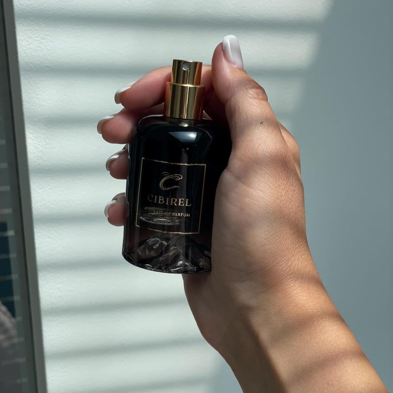 May include: A dark glass perfume bottle with a gold-colored spray nozzle and a black label featuring the gold text CIBIREL EXTRAIT DE PARFUM. The base of the bottle contains small, dark, irregular stones visible through the translucent glass.