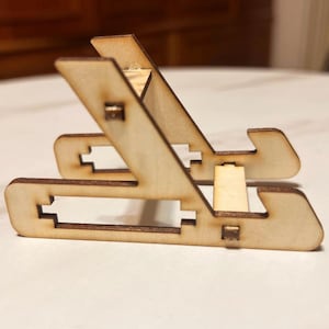 Puede incluir: Soporte de teléfono de madera natural. El soporte está hecho de piezas de madera contrachapada entrelazadas, con un diseño ranurado para sostener un teléfono en ángulo. El soporte es plegable y portátil.
