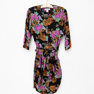 Puede incluir: Un vestido negro vintage con un estampado floral con flores moradas y rosas, diseños de cachemira y hojas verdes. El vestido tiene un cinturón con una hebilla circular y mangas tres cuartos. El vestido está colgado de una percha blanca.