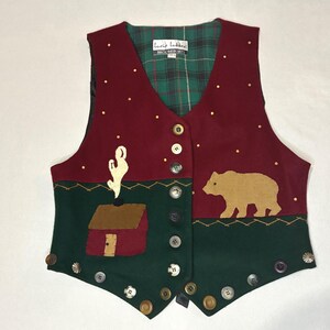 May include: A burgundy vest with a green and brown landscape design. The vest features a house, cactus, and bear applique, with gold-coloured dot accents and various button closures. The interior lining is a green and black plaid.