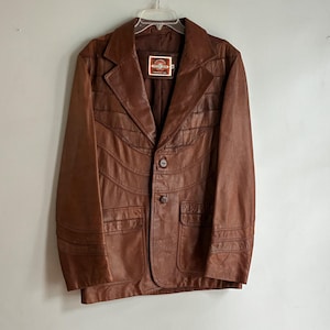 May include: A brown leather jacket featuring a notched lapel, two front buttons, and two lower flap pockets. The jacket displays decorative horizontal and curved stitched paneling across the chest and sleeves, with a label reading "Pioneer Wear" inside.