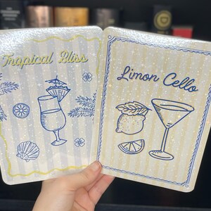 May include: Two rectangular coasters with a glittery, off-white background. One coaster reads "Tropical Bliss" and features a cocktail illustration. The other reads "Limon Cello" with a lemon and cocktail glass illustration. Both have blue and yellow accents.
