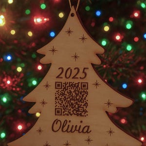 May include: A wooden Christmas tree ornament with the year 2025, a QR code, and the name "Olivia" engraved on it. The ornament is decorated with star shapes and hangs against a backdrop of colorful Christmas lights.