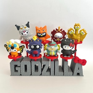 May include: A collection of eight Sanrio character figurines dressed as Godzilla monsters, displayed on a grey stone-textured base with the word GODZILLA embossed on the front and a red bow attached to the final letter A.
