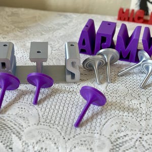 May include: Purple and silver decorative objects and the word "ARMY" in purple block letters are displayed on a white lace surface. The objects include silver tops and purple tops. The word "ARMY" is in large, block letters.