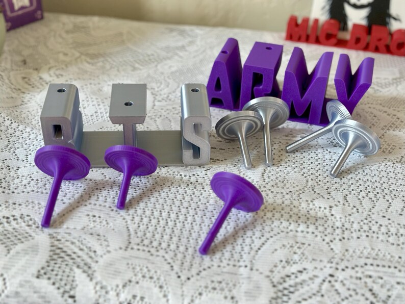 May include: Purple and silver decorative objects and the word "ARMY" in purple block letters are displayed on a white lace surface. The objects include silver tops and purple tops. The word "ARMY" is in large, block letters.