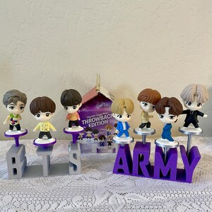 May include: A collection of small figurines of cartoon characters, each standing on a pedestal. The figurines are arranged on a table with the letters "BTS" and "ARMY" in front of them. A box with the words "THROWBACK EDITION" is visible.