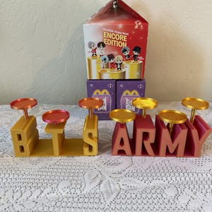 May include: A display featuring the words "BTS" and "ARMY" in gold and red, each letter holding a small, round tray. A Happy Meal box with "ENCORE EDITION" text and character illustrations sits behind the letters.
