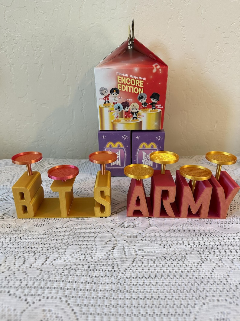 May include: A display featuring the words "BTS" and "ARMY" in gold and red, each letter holding a small, round tray. A Happy Meal box with "ENCORE EDITION" text and character illustrations sits behind the letters.