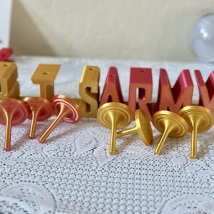 May include: Gold and red-orange 3D-printed letters spelling "BTS ARMY" with several gold and red-orange tops. The letters are on a white lace tablecloth. A light stick is visible in the background.
