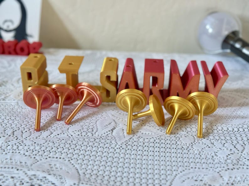 May include: Gold and red-orange 3D-printed letters spelling "BTS ARMY" with several gold and red-orange tops. The letters are on a white lace tablecloth. A light stick is visible in the background.