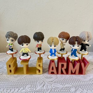 May include: A collection of seven small figurines of people, each standing on a small platform. The figurines are arranged above the letters "BTS ARMY" in gold and red. The background is a light beige color.