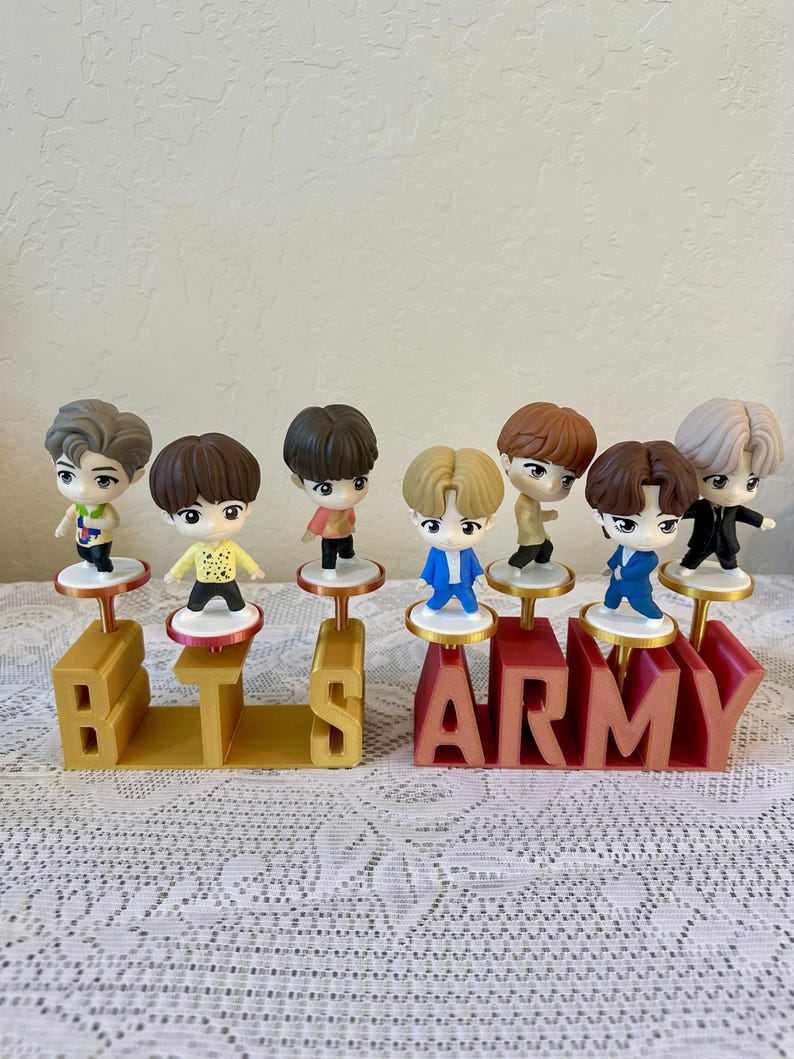 May include: A collection of seven small figurines of people, each standing on a small platform. The figurines are arranged above the letters "BTS ARMY" in gold and red. The background is a light beige color.