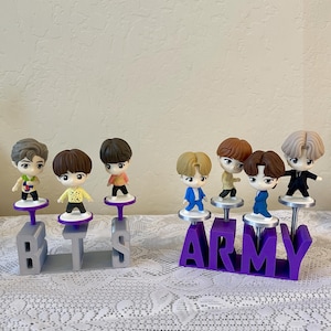 May include: A collection of small figurines of cartoon characters, each standing on a purple or gray base. The bases spell out "BTS" and "ARMY". The figurines have different hair colors and outfits.