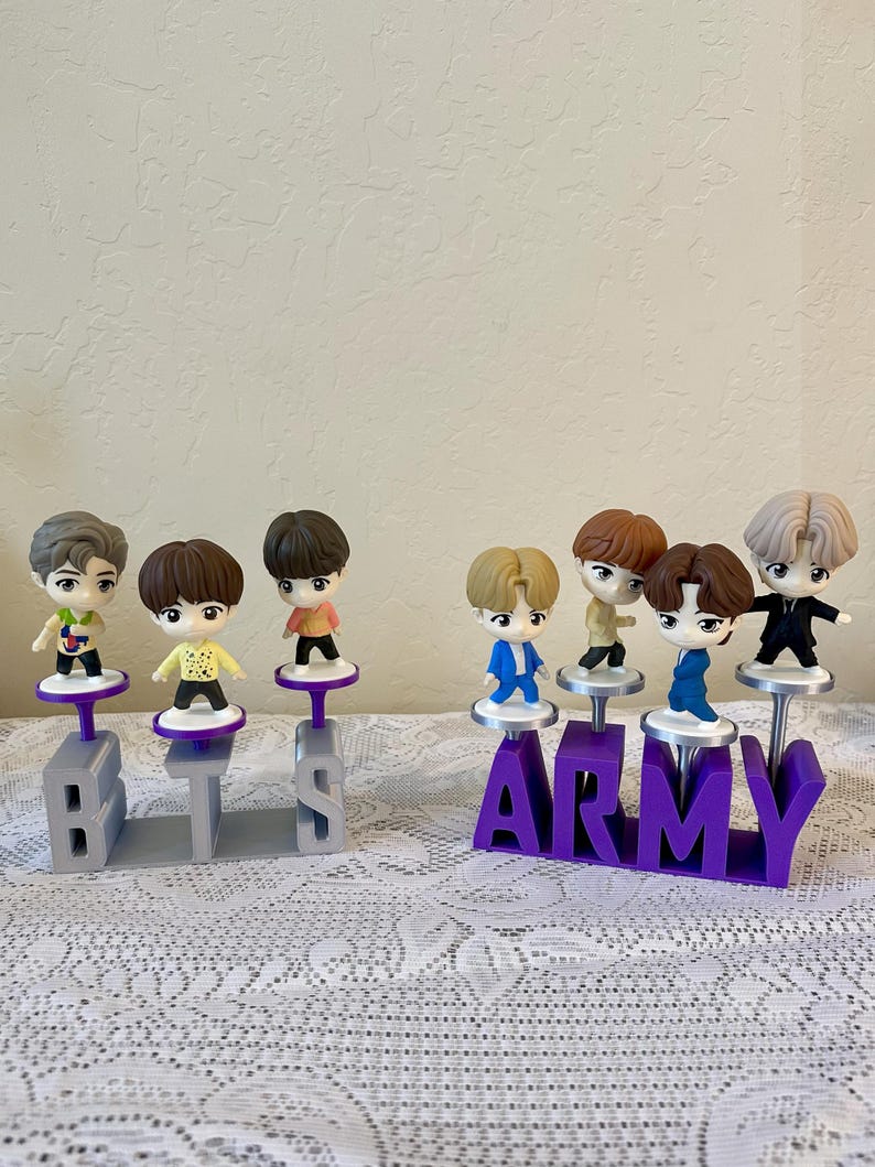 May include: A collection of small figurines of cartoon characters, each standing on a purple or gray base. The bases spell out "BTS" and "ARMY". The figurines have different hair colors and outfits.