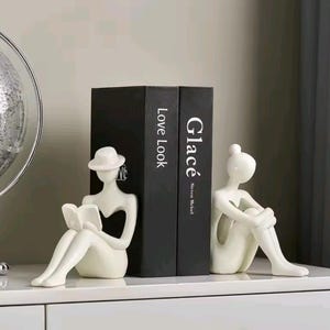 Puede incluir: Soportes para libros de cerámica blanca con figuras abstractas. Una figura está sentada leyendo un libro y lleva un sombrero. La otra figura está sentada con los brazos alrededor de las rodillas. Libros negros con texto blanco entre las figuras.