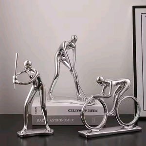 Puede incluir: Tres esculturas plateadas que representan figuras atléticas. Una figura balancea un bate de béisbol, otra un palo de golf y la tercera monta en bicicleta. Las esculturas se exhiben sobre una superficie oscura.